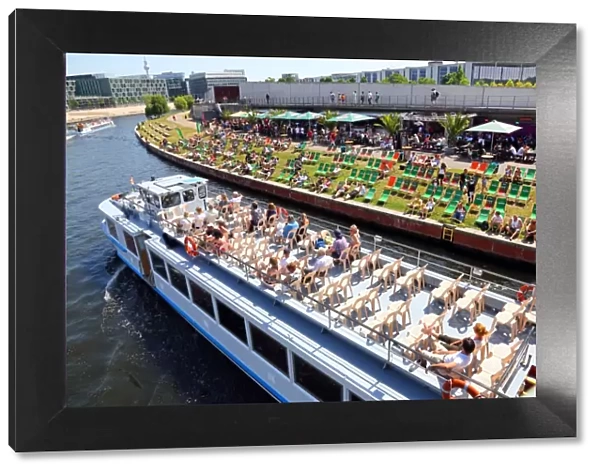 Artificial beach with deckchairs beside the River Spree with a tourist sightseeing boat in Berlin, Germany